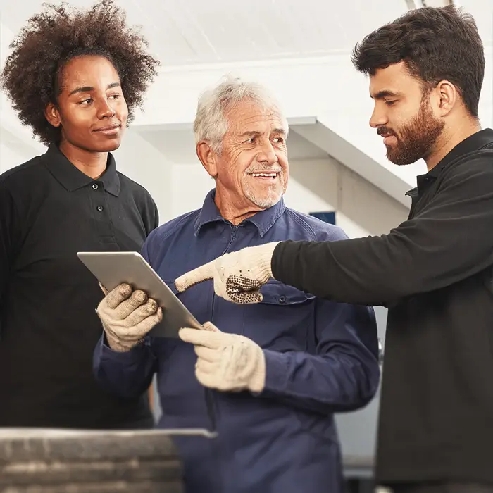 Ein älterer Herr im blauen Hemd und mit einem Tablet in der Hand ist im Gespräch mit zwei jüngeren Lehrlingen, die eine Ausbildung in der Automobil-Branche machen.