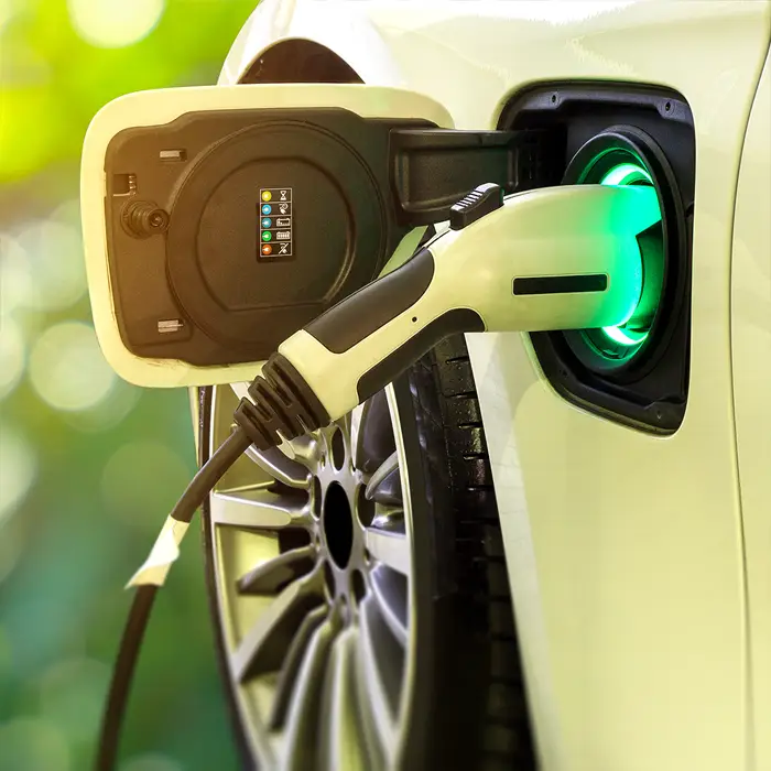 Ein beigefarbenes E-Auto ist mit einem Ladestecker verbunden, um die Batterien aufzuladen. Die Steckerbuchse leuchtet innen grün.