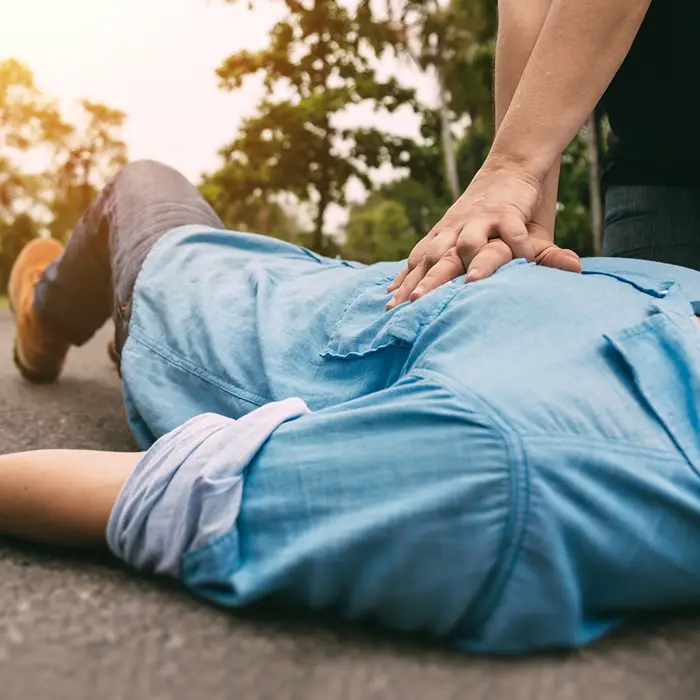 Auf einer Straße liegt ein lebloser Körper. Eine zweite Person gibt Erste Hilfe und macht mit seinen beiden Händen eine Herzdruckmassage.