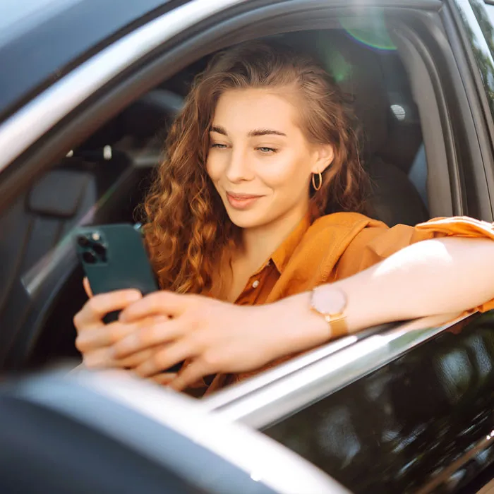 Influencer sind affin für Social Media. Hier sitzt eine junge Frau mit braunen lockigen Haaren hinter dem Steuer eines Fahrzeuges und bedient mit beiden Händen ihr Smartphone. Die Fensterscheibe ist geöffnet und sie lehnt sich mit ihrem linken Arm heraus.