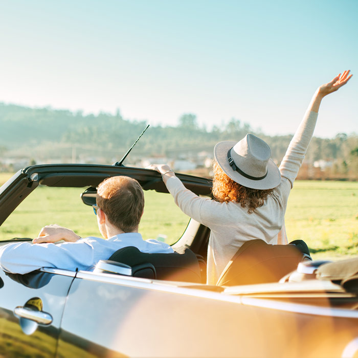 Leidenschaft Cabrios: Ein Mann und eine Frau fahren im offenen Cabrio durch eine schöne Landschaft. Die Frau mit Sonnenhut lässt ihren hoch erhobenen Arm durch den Wind streifen. Die Landschaft spiegelt sich auf dem Autolack.