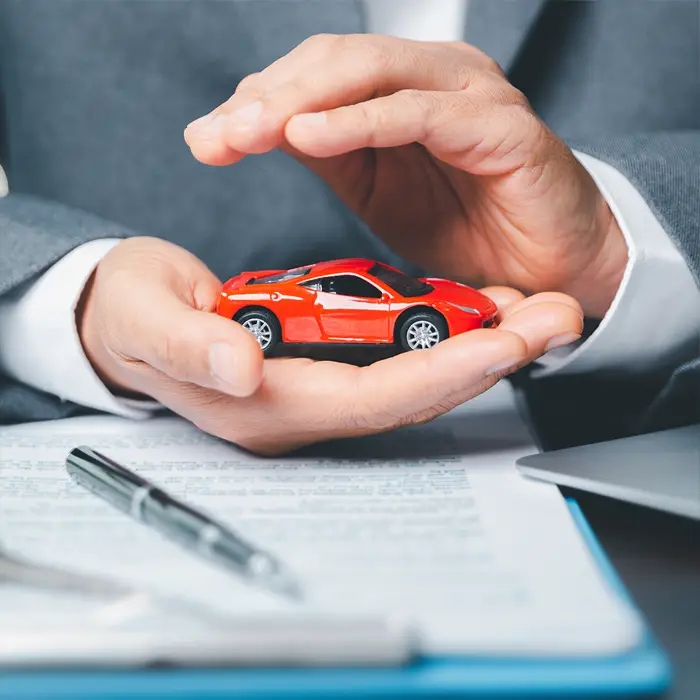 Ein Mann von der Versicherung hält in seiner einen Hand ein rotes Spielzeugauto. Seine zweite Hand hält er schützend über das Auto, um es symbolisch vor Autodiebstahl zu bewahren.