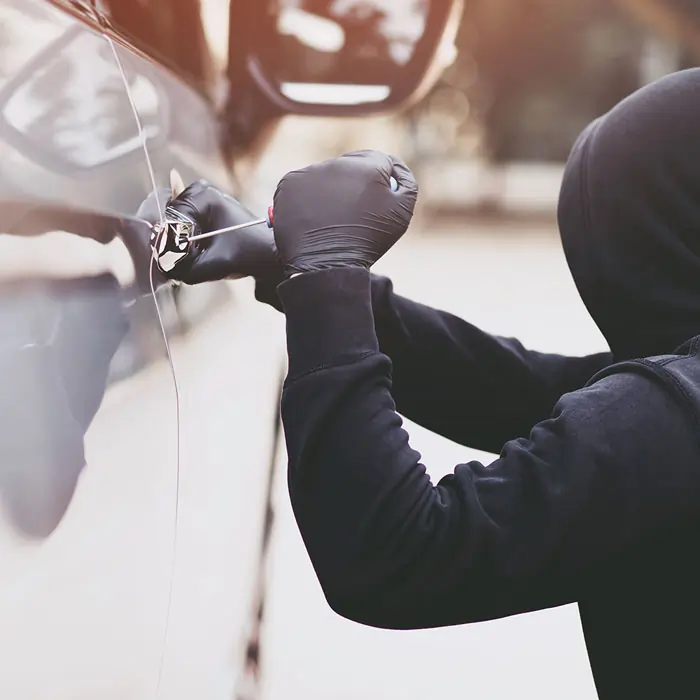 Ein schwarz gekleideter und vermummter Autodieb versucht mit einem spitzen Werkzeug ähnlich eines Schraubendrehers das Schloss einer Autotür zu knacken.